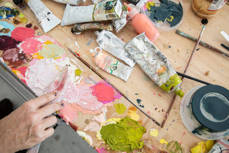 Artistic workspace with paint tubes, brushes, and a palette of colorful paint on a wooden table.
